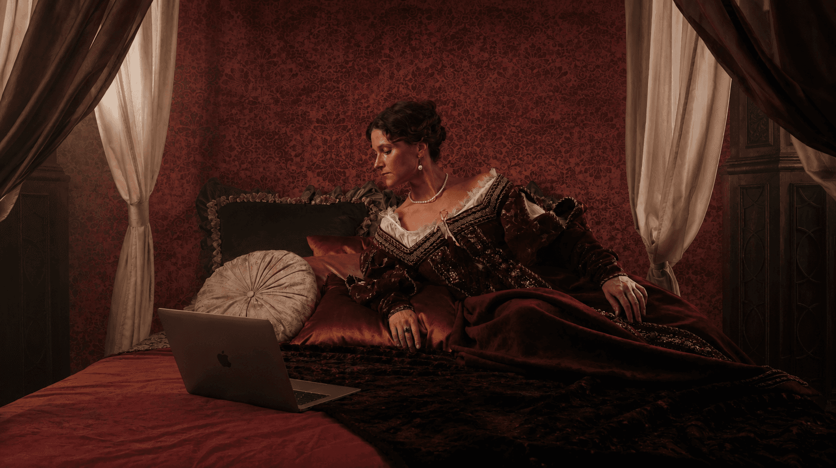 A figure in period dress reclining on a bed with a laptop